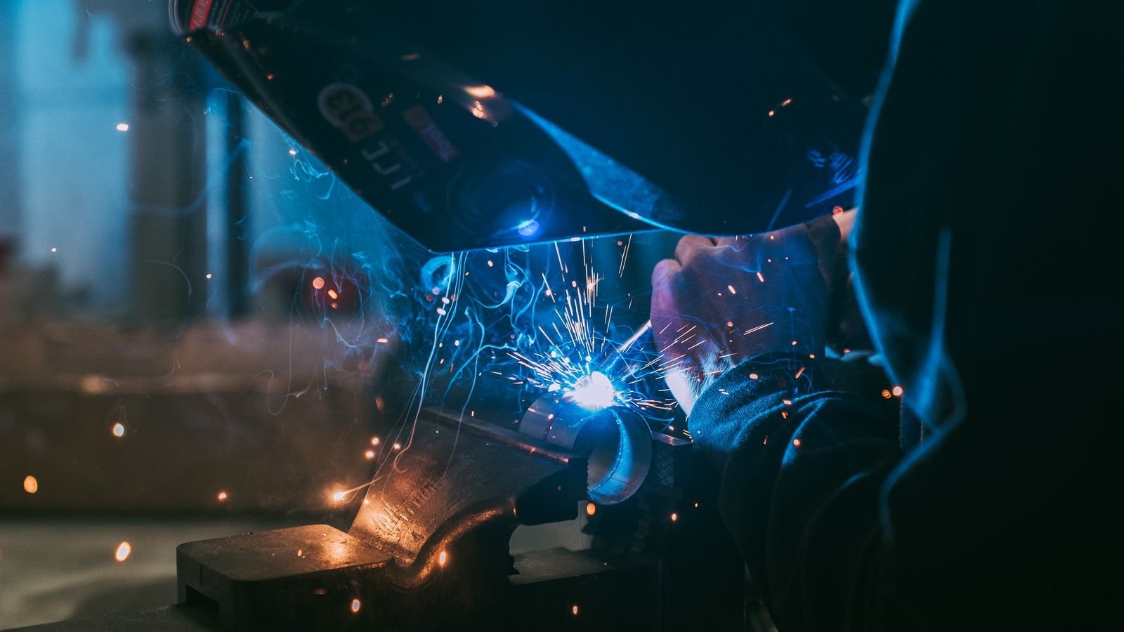 A welder laying a bead on steel, arc light and sparks filling the frame.